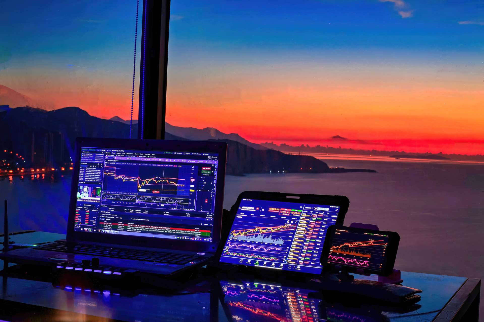 A photo of a laptop, a table, and a phone on a table, showing charts and trading platforms